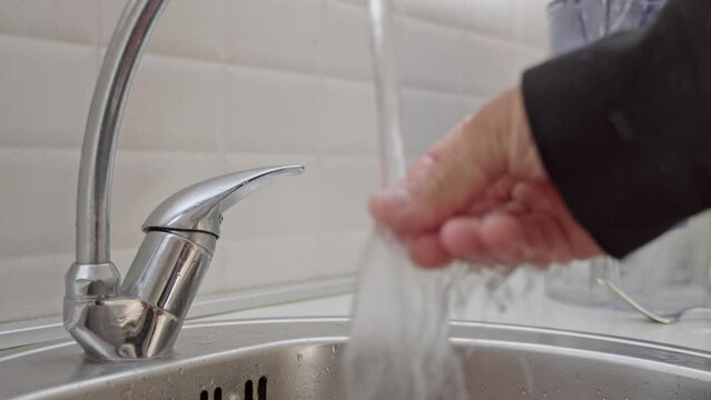 A man in a black jacket opens a tap of water to rinse his hands in the sink against the background of a white wall, A man washes his hand in the sink. Close-up
