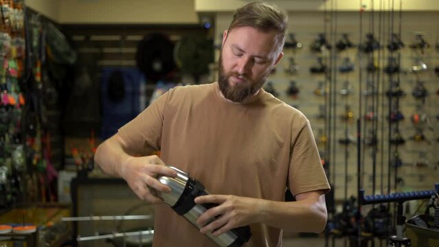 bearded man in a fishing tackle shop getting ready for a fishing trip. Chooses a thermos for a trip is determined by the size compares large and small