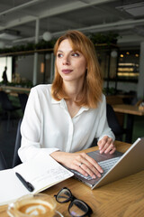 White young woman in a white shirt with silver watches sitting at the wooden table and drinking coffee from a transparent cup