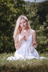 Fototapeta na wymiar young blond girl sitting on the grass and smiling, a fashion model, a woman in a white dress