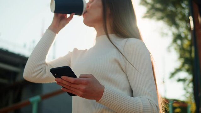 Woman using smartphone outdoor. Young female student texting at phone, walking in street, drinking coffee. Communicating with family and friends online via internet messages.