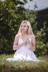 Fototapeta na wymiar young blond girl sitting on the grass and smiling, a fashion model, a woman in a white dress