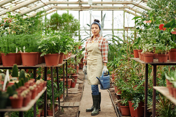 Fototapeta na wymiar Horizontal long shot of Hispanic woman wearing apron and rubber boots holding watering can standing in greenhouse looking at camera