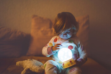 Cute little toddler girl playing with colorful night light lamp before going to bed. Sleepy tired...