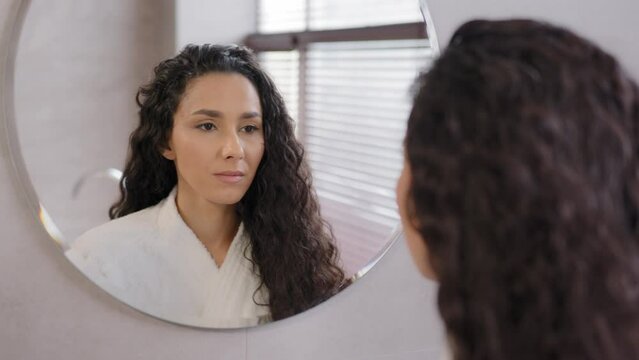Close-up serious pensive young hispanic woman looking in mirror in bathroom contemplating future cute curly brunette girl in bathrobe admires reflection natural beauty daily morning routine concept