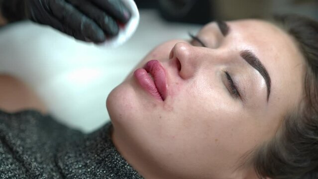 High angle view face of young slim woman in beauty salon with beautician removing blood and red pigment leftovers from lips. Beautiful Caucasian confident client doing permanent lip tattooing