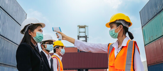 asian business woman and team wearing face mask checking fever by thermometer before entering work in logistic cargo warehouse at manufacturing to protect from covid or coronavirus.
