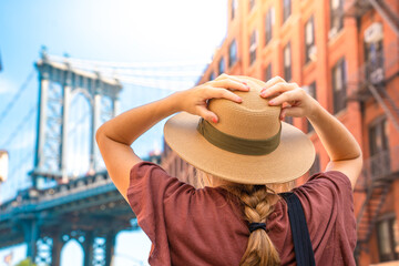 Fashionable tourist woman visiting New York City wearing hat and stylish street style outfit. Young girl amazed by the beautiful city. Manhattan bridge view on summertime.