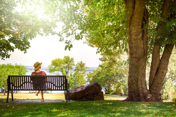 Back view of young attractive woman sitting and relaxing on a bench outdoors in beautiful nature park. - obrazy, fototapety, plakaty