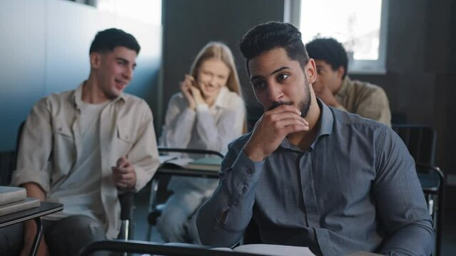 Sad frustrated arab student sitting in class at desk alone suffering from mistreatment by classmates racial discrimination feels victim of bullying violent students bully ridicule gossip behind back