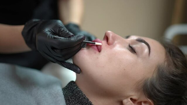 Beautician tattoo master covering tattooed female lips with cream in beauty salon. Side view portrait of gorgeous Caucasian woman after permanent makeup tattooing receiving aftercare