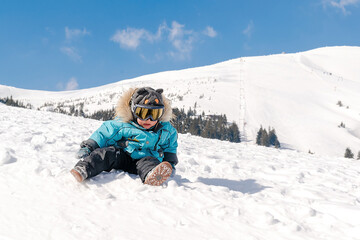 Fototapeta na wymiar Little cute baby kid child boy in winter overalls,ski mask,glasses goggles sitting on snow at winter holiday, in ski resort in Carpathian high mountains outdoor nature landscape, Ukraine, Europe