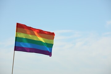 Bright LGBT flag against blue sky with clouds, space for text