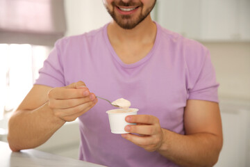 Happy young man with tasty yogurt at table in kitchen, closeup