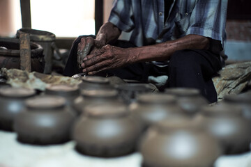 Selective focused on the dirty wrinkled skin hands of old man molding the clay work on the spinning wheel for making the jar with blurred group of clay jars in foreground