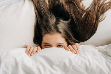 In the morning, a young woman peeps out from under the blanket lying on the bed.
