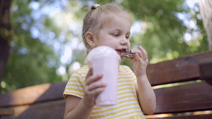 ittle girl eats a colorful gingerbread and holds a milkshake in her hand. Close-up of cute child girl sitting on park bench and eating cookies with a milkshake.