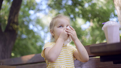 Little girl wipes her face with a paper napkin after eating, there is a milkshake on the table. Close-up of cute child girl sitting on park bench and wiping her face with a paper napkin.