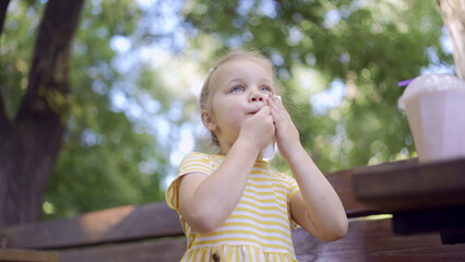 Naklejka premium Little girl wipes her face with a paper napkin after eating, there is a milkshake on the table. Close-up of cute child girl sitting on park bench and wiping her face with a paper napkin.