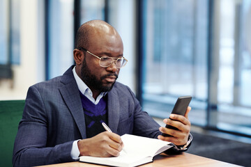 African businessman in suit looking at the screen of mobile phone writing the plans in his...