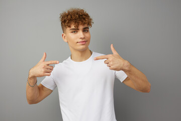 a curly-haired young man stands in a white T-shirt on a studio background, isolated from the background and pleasantly smiling, pointing at himself with his index fingers