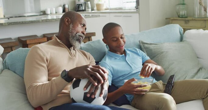 Video of happy african american father and son eating chips and watching tv together