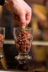A close up portrait of a hand putting a leaf of mint on top of some sweet and tasty brown chocolate mousse in a glass cup ready to be served and eaten.