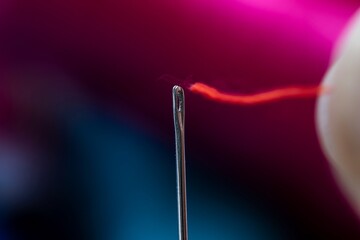 A portrait of a metal sewing needle with someone trying to put a red thread through the eye of the piece of sewing equipment.