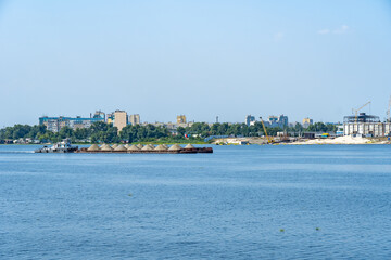 Fototapeta premium Tugboat moves barge with sand along the picturesque river. Cargoship to transport along the waterway. Large heaps of sand are transported by ship. Infrastructure big city.