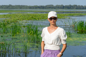 Portrait of woman on the background overgrown river. She is wearing white blouse and cap. Slowly flowing river is overgrown with yellow water lilies. Ecological problem.