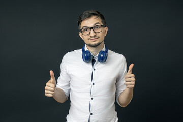 young man in glasses with headphones around his neck shows two thumbs up and stands on a black background