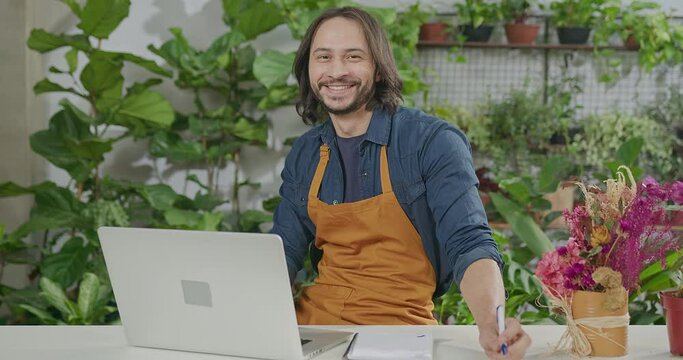 Portrait of a male employee wearing apron in front of laptop smiling at camera receiving online orders at small business flower shop retail store. Happy worker in front of computer