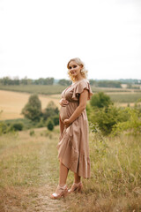 A pregnant blonde woman in a dress touches her belly in a wheat field at sunset.