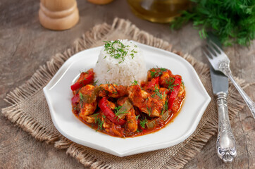 Chicken saute. Braised Chicken breast in Tomato Sauce mixed with onion and colorful bell pepper served in white plate with cooked rice. Wooden background, selective focus.
