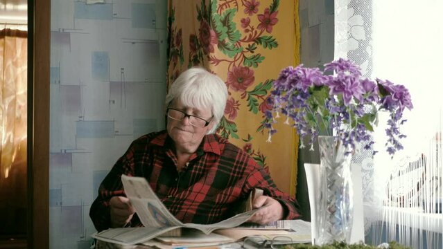 Portrait of an old woman in glasses reading a newspaper while sitting at a table at home