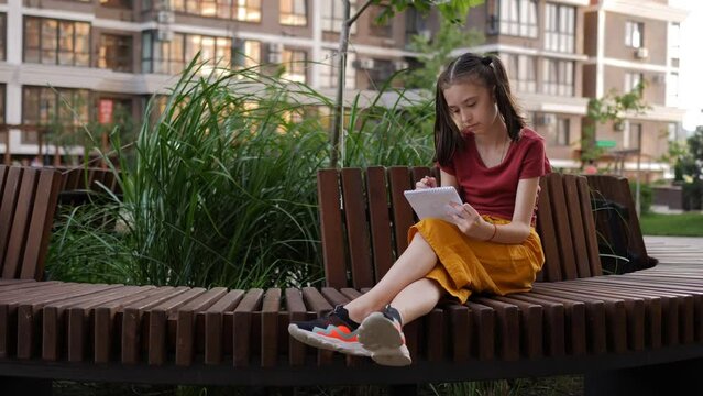 A creative little girl draws in a notebook on the street sitting on a bench.