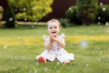 happy smiling baby girl in white dress is sitting outdoors on green grass on summer day. child...