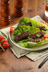 Traditional south european skinless sausages cevapcici made of ground meat and spices on lettuce leaves with thyme and tomatoes on white plate on kitchen towel, black silverware