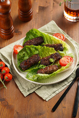 Traditional south european skinless sausages cevapcici made of ground meat and spices on lettuce leaves with thyme and tomatoes on white plate on kitchen towel, black silverware
