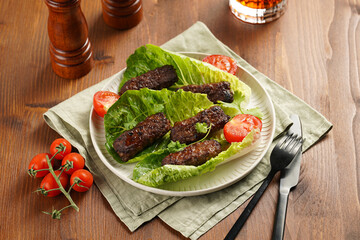Traditional south european skinless sausages cevapcici made of ground meat and spices on lettuce leaves with thyme and tomatoes on white plate on kitchen towel, black silverware