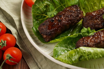 Traditional south european skinless sausages cevapcici made of ground meat and spices on lettuce leaves with thyme and tomatoes on white plate on kitchen towel, black silverware
