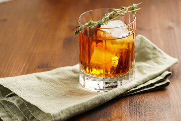 a tumbler glass with brown alcoholic drink, thyme and ice cubes on linen napkin - whisky, rum or cognac - on wooden table, rural scene