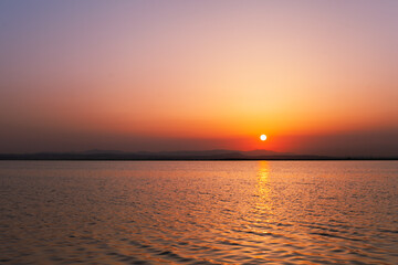 Colorful orange, yellow and purple sunset over water of lake with sun setting in the water. Natural park at sunset with distant silhouettes of mountains. Sunset landscape view from a boat. Traveling 