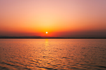 Colorful orange, yellow and red sunset over the water of lake with sun setting in the water. Natural park at sunset with distant silhouettes of mountains. Sunset landscape view from a boat. Traveling 