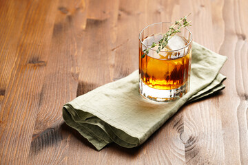 a tumbler glass with brown alcoholic drink, thyme and ice cubes on linen napkin - whisky, rum or cognac - on wooden table, rural scene
