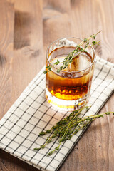 a tumbler glass with brown alcoholic drink, thyme and ice cubes on linen napkin - whisky, rum or...