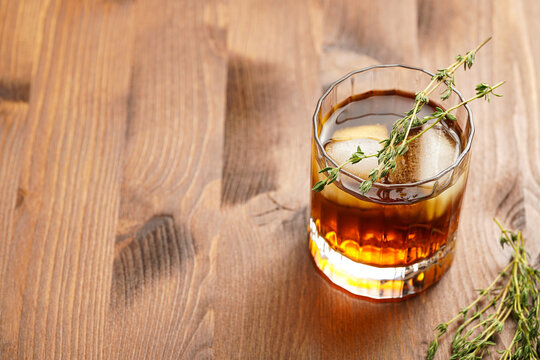 a tumbler glass with brown alcoholic drink, thyme and ice cubes - whisky, rum or cognac - on wooden table, rural scene