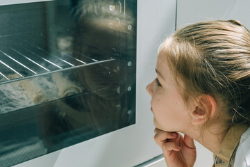 Fototapeta na wymiar Girl baking homemade cookies and looking inside oven through glass