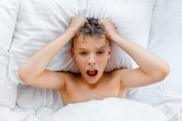 Worried awake teen boy in a bed is shocked because it is too late. Kid lies on the pillow and clutched his head in horror because he is overslept