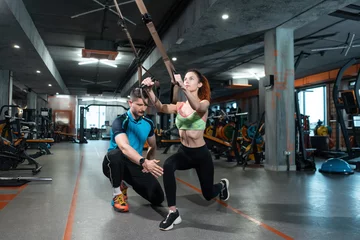 Tuinposter young female exercising and has workout with trx and personal trainer in gym © goami